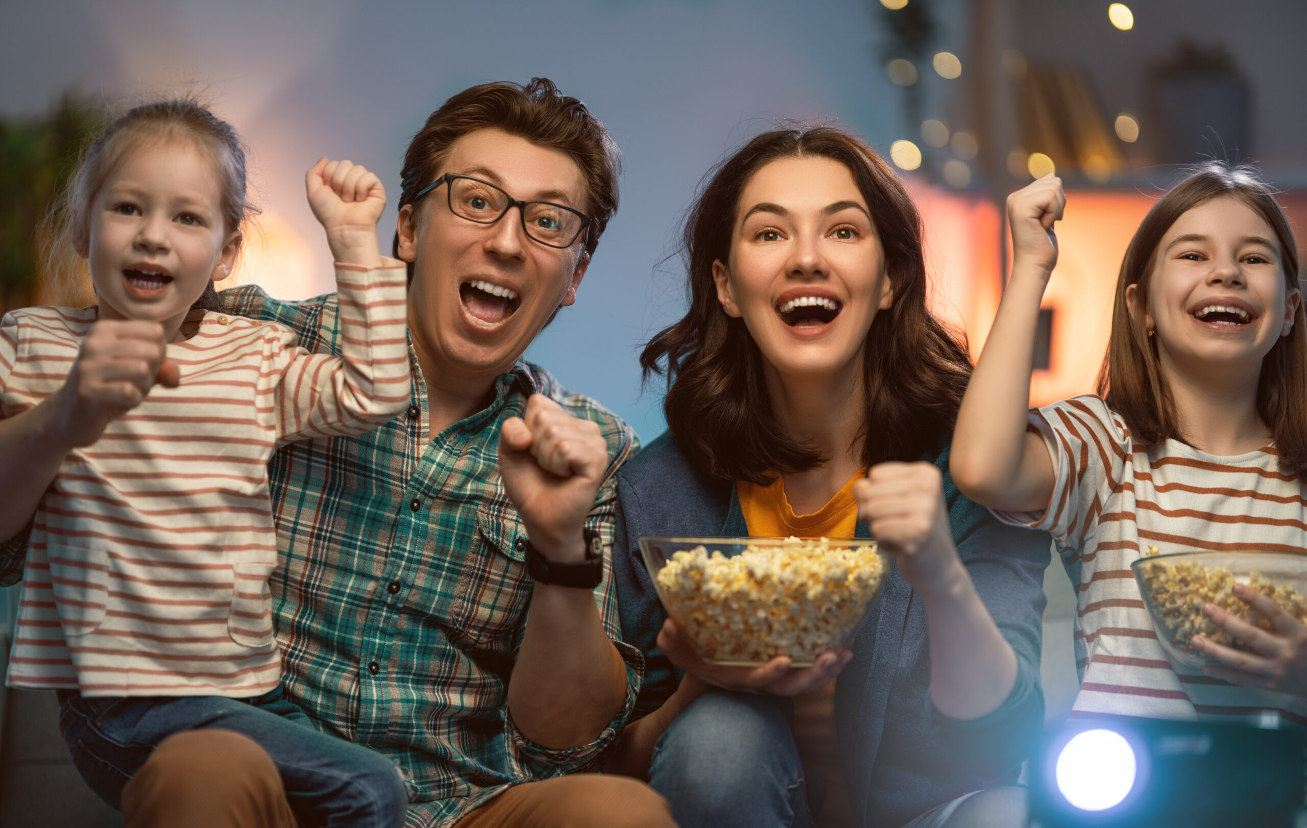 Happy family watching projector, TV, movies with popcorn in the evening at home. Mother, father and daughters spending time together.